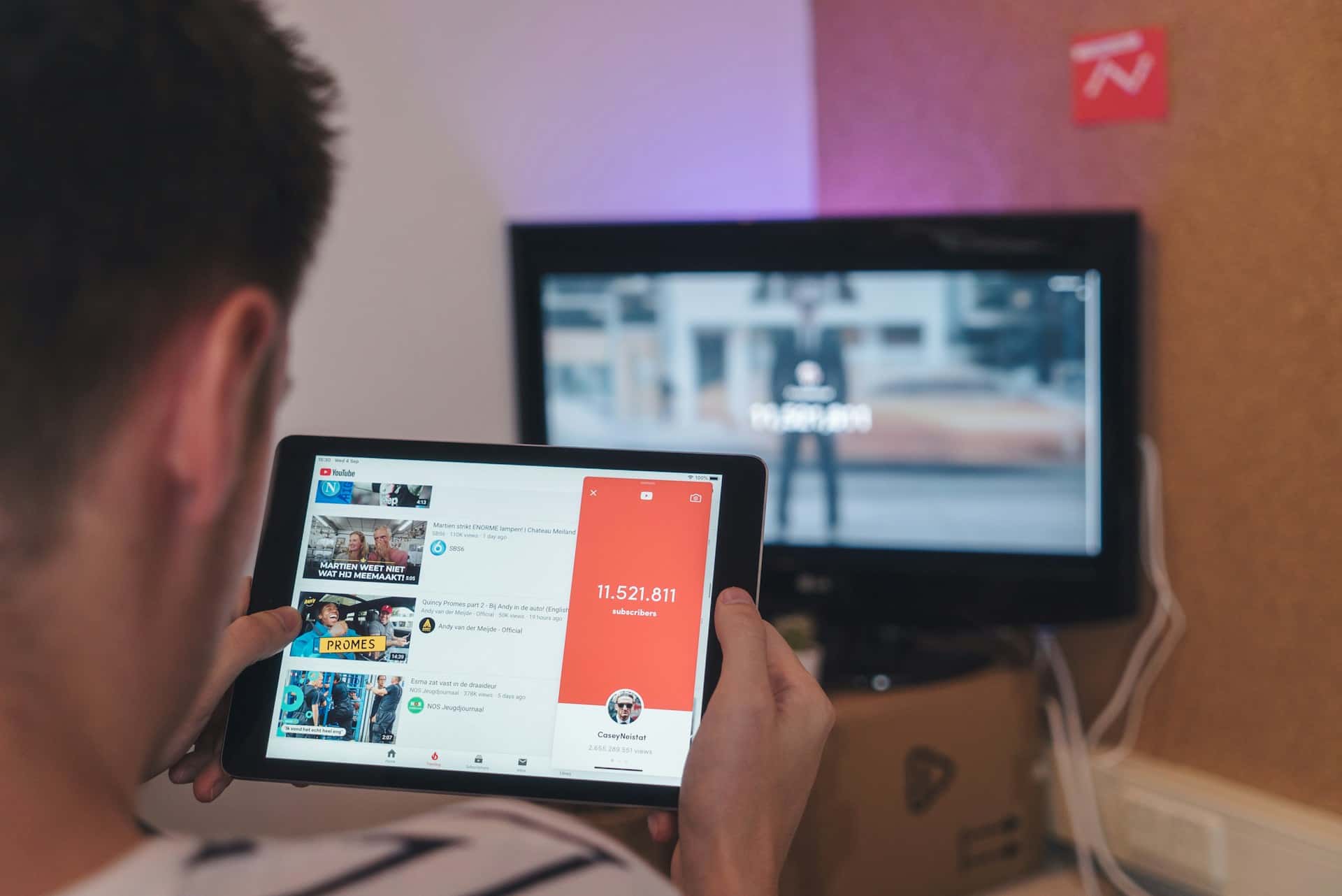 Vue par-dessus l'épaule d'une personne consultant YouTube sur une tab--lette, avec une télévision en arrière-plan. L'interface YouTube affiche une liste de vidéos et un compteur d'abonnés dépassant 11 millions