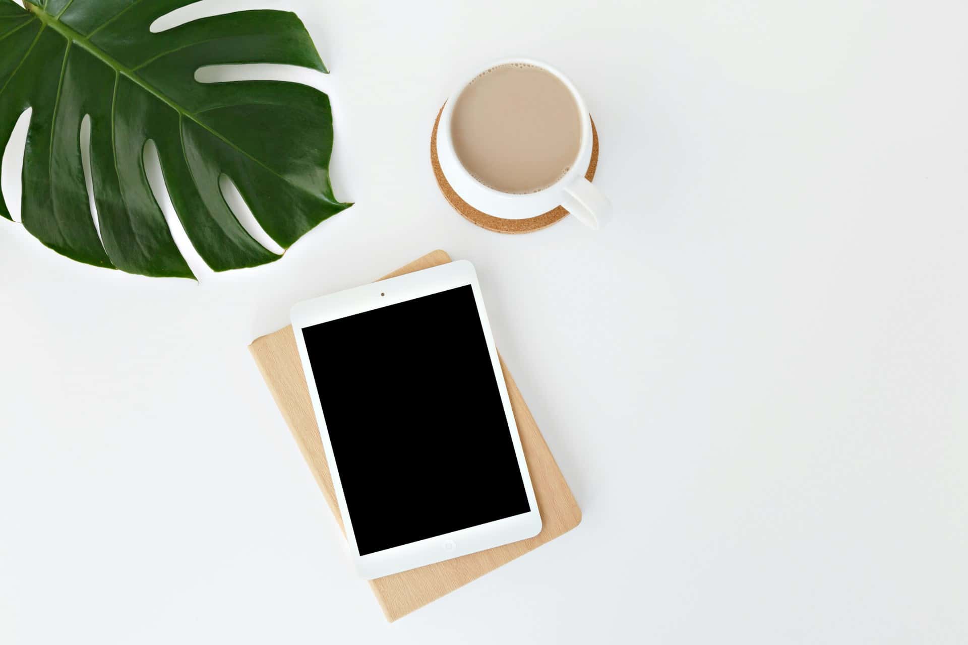 Vue en plongée d'un espace de travail minimaliste sur fond blanc avec une tablette à écran noir, une tasse de café au lait sur un dessous de verre en liège, et une grande feuille de Monstera verte