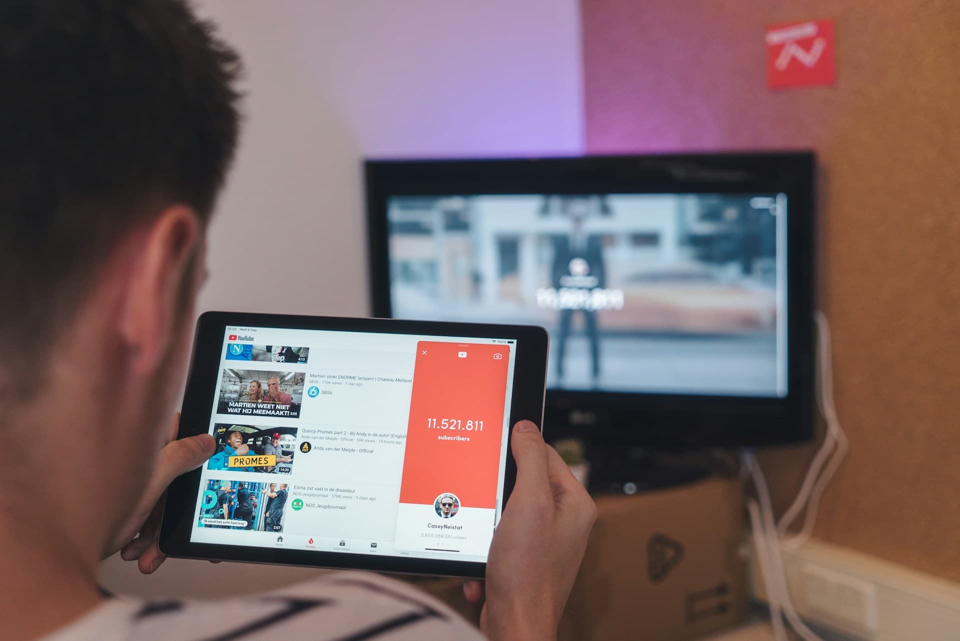 Vue par-dessus l'épaule d'une personne consultant YouTube sur une tab--lette, avec une télévision en arrière-plan. L'interface YouTube affiche une liste de vidéos et un compteur d'abonnés dépassant 11 millions