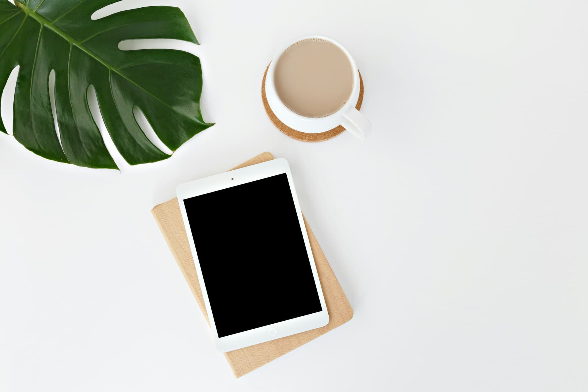 Vue en plongée d'un espace de travail minimaliste sur fond blanc avec une tablette à écran noir, une tasse de café au lait sur un dessous de verre en liège, et une grande feuille de Monstera verte