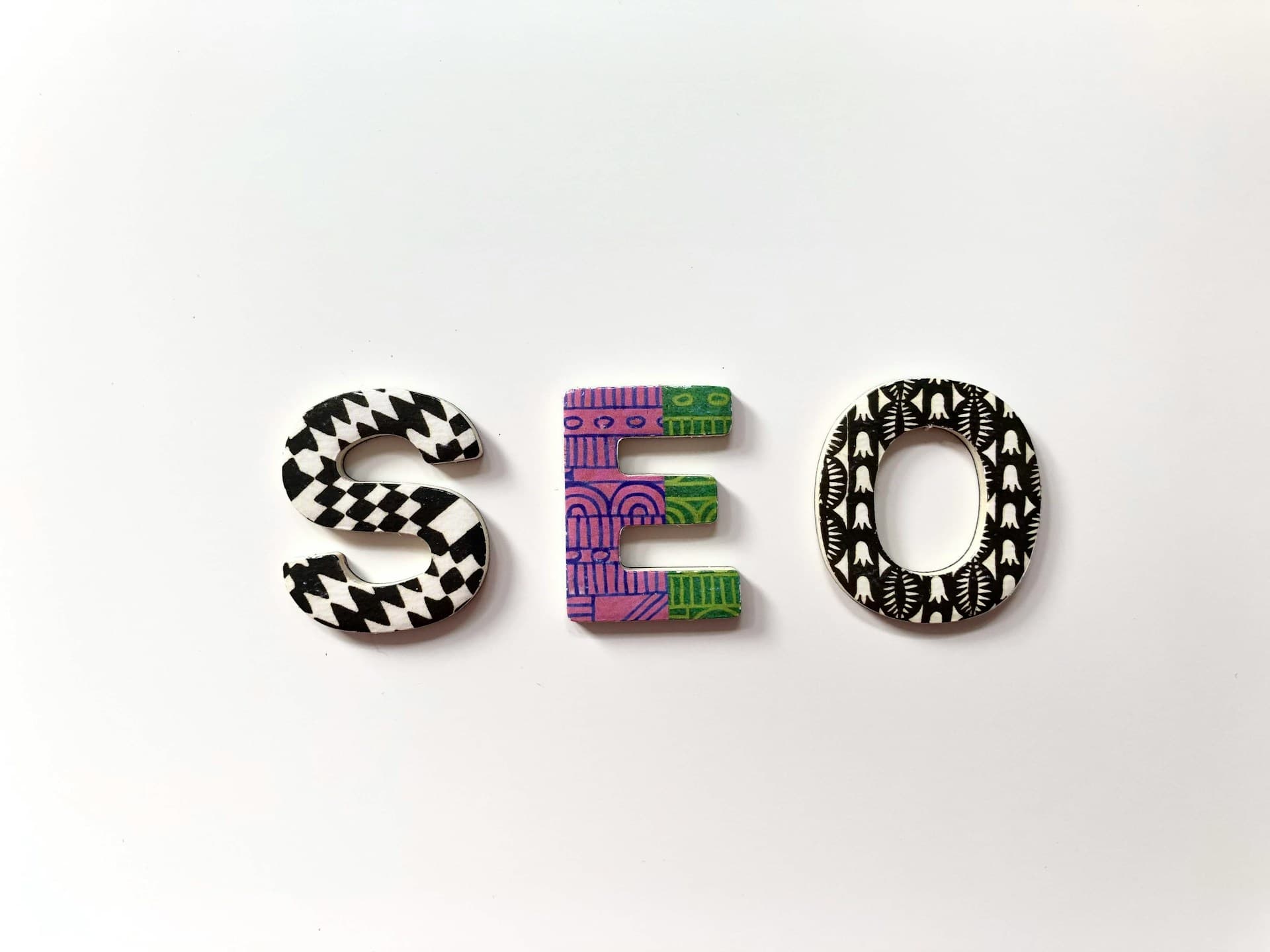 Le mot 'SEO' composé de trois lettres décoratives sur fond blanc - 'S' avec motif damier noir et blanc, 'E' avec motifs géométriques violet et vert, et 'O' avec motif floral noir et blanc