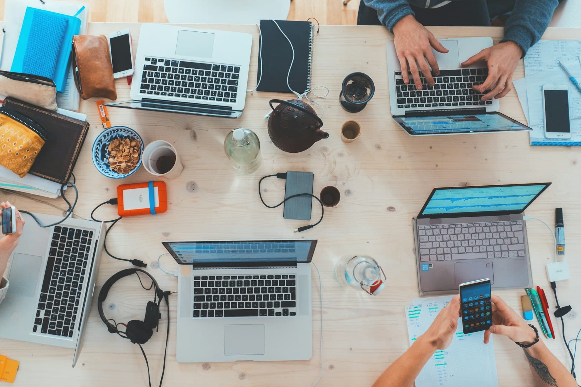 Vue aérienne d'un espace de travail collaboratif avec plusieurs ordinateurs portables, des accessoires tech comme des casques et disques durs externes, des tasses de café et des en-cas sur une table en bois clair